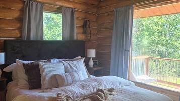 A wooden cabin bedroom with a bed, pillows, a blanket, a nightstand, a lamp, and a view of the outdoors.