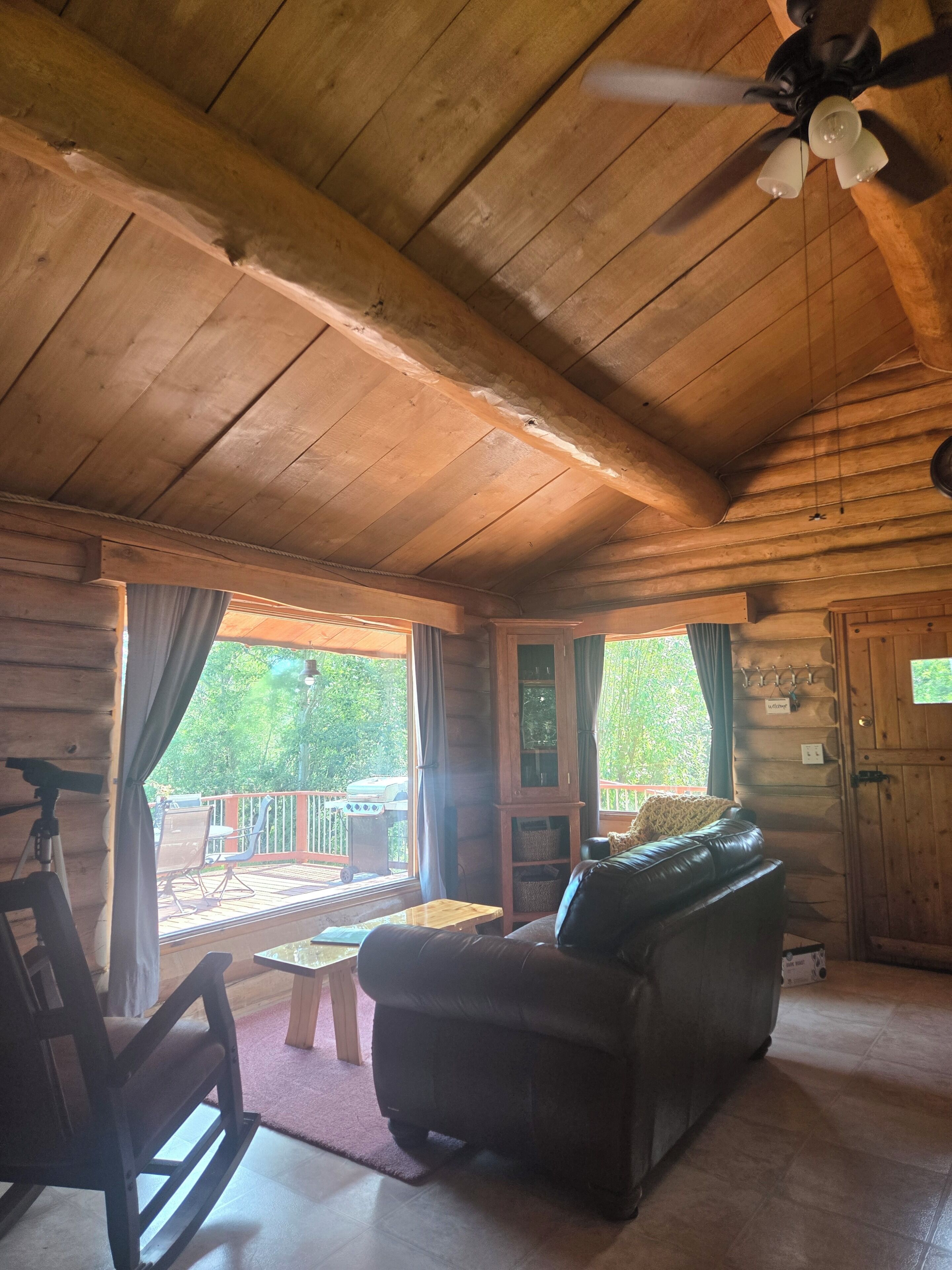 A wooden cabin interior with a ceiling fan, a leather sofa, a rocking chair, and a view of the outdoors.