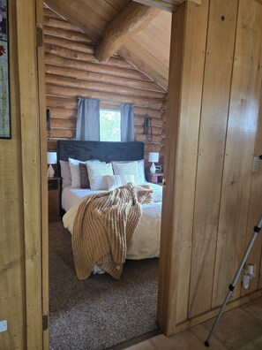 A wooden cabin bedroom with a bed, a nightstand, a lamp, and a window with curtains.