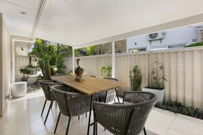 Une terrasse couverte avec une table en bois et des chaises en osier, entourée de plantes en pot et d’une clôture blanche.