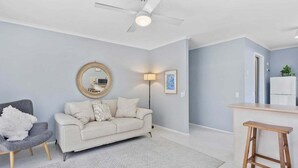 A living room with a sofa, a chair, a coffee table, a ceiling fan, and a kitchen area with a counter and stools. - 2BR Paradise at Riviera (Gold Coast)