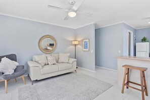 Ein Wohnzimmer mit Sofa, Sessel, Couchtisch, Deckenventilator und einer Küchenzeile mit einer Arbeitsplatte und Hockern.
