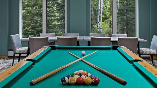A pool table with balls and cues, surrounded by chairs and tables, with large windows and a view of trees. -  Spacious 2-bedroom condo in Magical Canmore with fitness room and AC (Canmore)