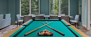 Une table de billard avec des billes et des queues, entourée de chaises et de tables, avec de grandes fenêtres donnant sur un paysage arboré.