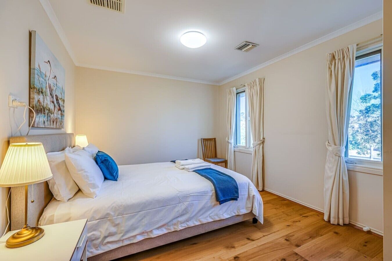 Un dormitorio con cama, lámpara de noche, ventana con cortinas y un cuadro en la pared.
