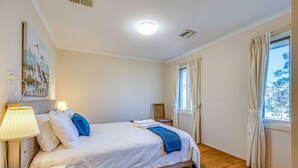 Un dormitorio con cama, lámpara de noche, ventana con cortinas y un cuadro en la pared.