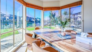 Un comedor moderno con una gran mesa de madera, sillas con cojines y ventanas amplias que ofrecen vistas a una zona residencial y una masa de agua.