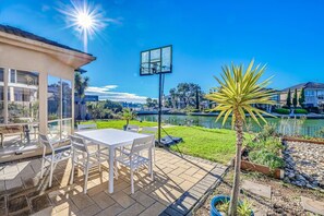 Eine Terrasse mit Tisch und Stühlen, ein Basketballkorb und Blick auf einen See mit Gebäuden im Hintergrund.