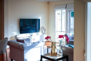 Ein Wohnzimmer mit Flachbildfernseher, weißen Möbeln, einer dekorativen Vase und Blick durch große Fenster.