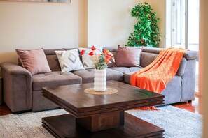 Ein Wohnzimmer mit einem dunklen Holztisch, einem grauen Sofa mit gemusterten Kissen und einer Zimmerpflanze im Hintergrund.