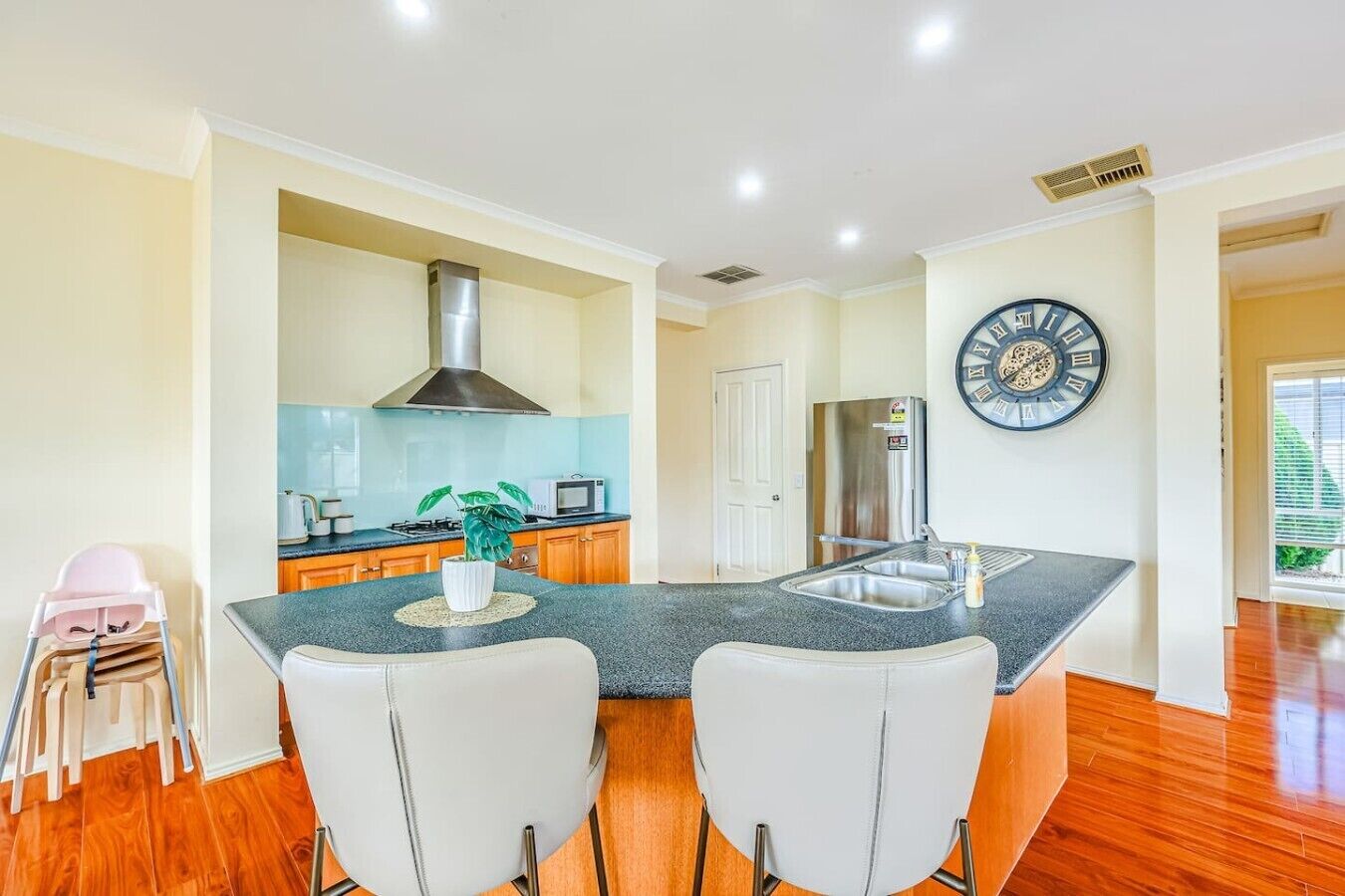 A kitchen with a central island, two white chairs, a stainless steel refrigerator, a microwave, and a clock on the wall.
