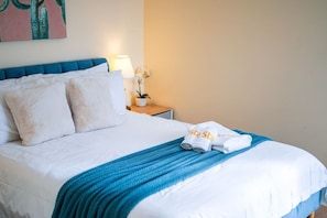 Ein ordentlich bezogenes Bett mit weißer Bettwäsche und blauem Akzent, ein kleiner Nachttisch mit Lampe und Topfpflanze sowie eine Wand mit einem farbenfrohen abstrakten Gemälde.