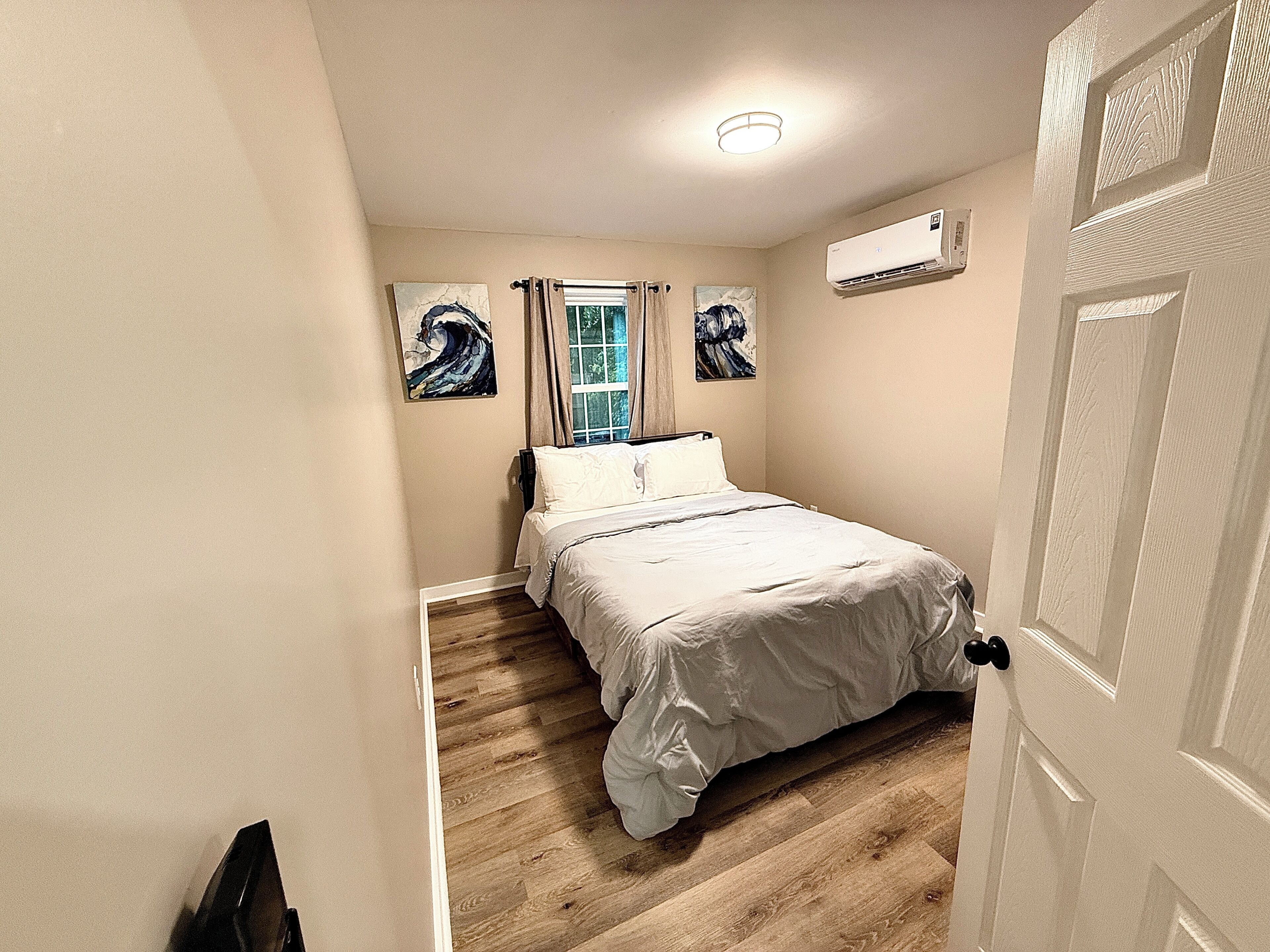 A bedroom with a bed, a window with curtains, a painting on the wall, and an air conditioning unit.