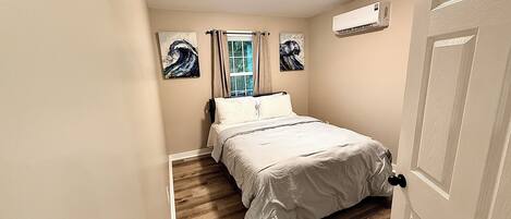 Un dormitorio con cama, ventana con cortinas, un cuadro en la pared y un equipo de aire acondicionado.