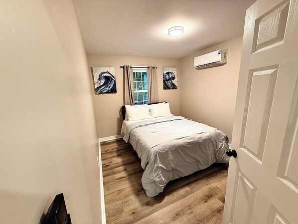 A bedroom with a bed, a window with curtains, a painting on the wall, and an air conditioning unit.