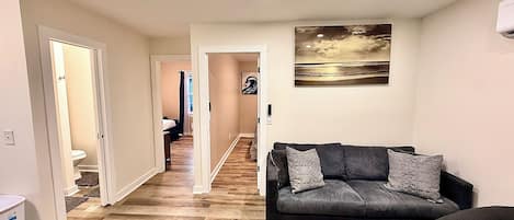 A hallway leading to a bedroom with a bed, a living room with a sofa and a coffee table, and a painting on the wall.