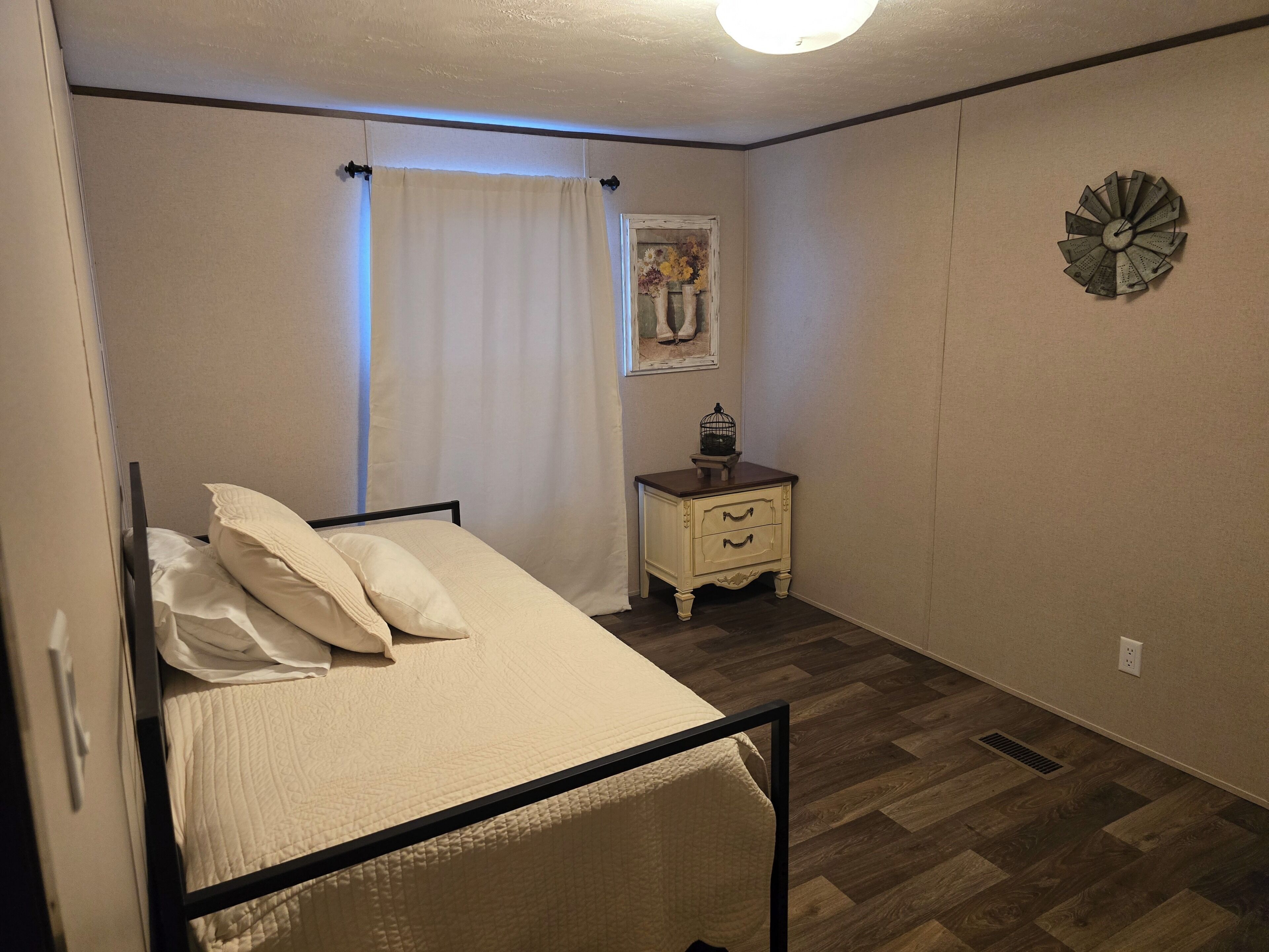 A bedroom with a bed, a nightstand, a wall clock, and a window with curtains.