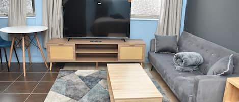 A modern living room with a flat-screen TV, a grey sofa, a wooden coffee table, and a small round table.