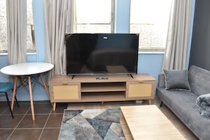 Un salon avec un téléviseur à écran plat posé sur un meuble télé en bois, une table ronde blanche, un canapé gris et un tapis à motifs.