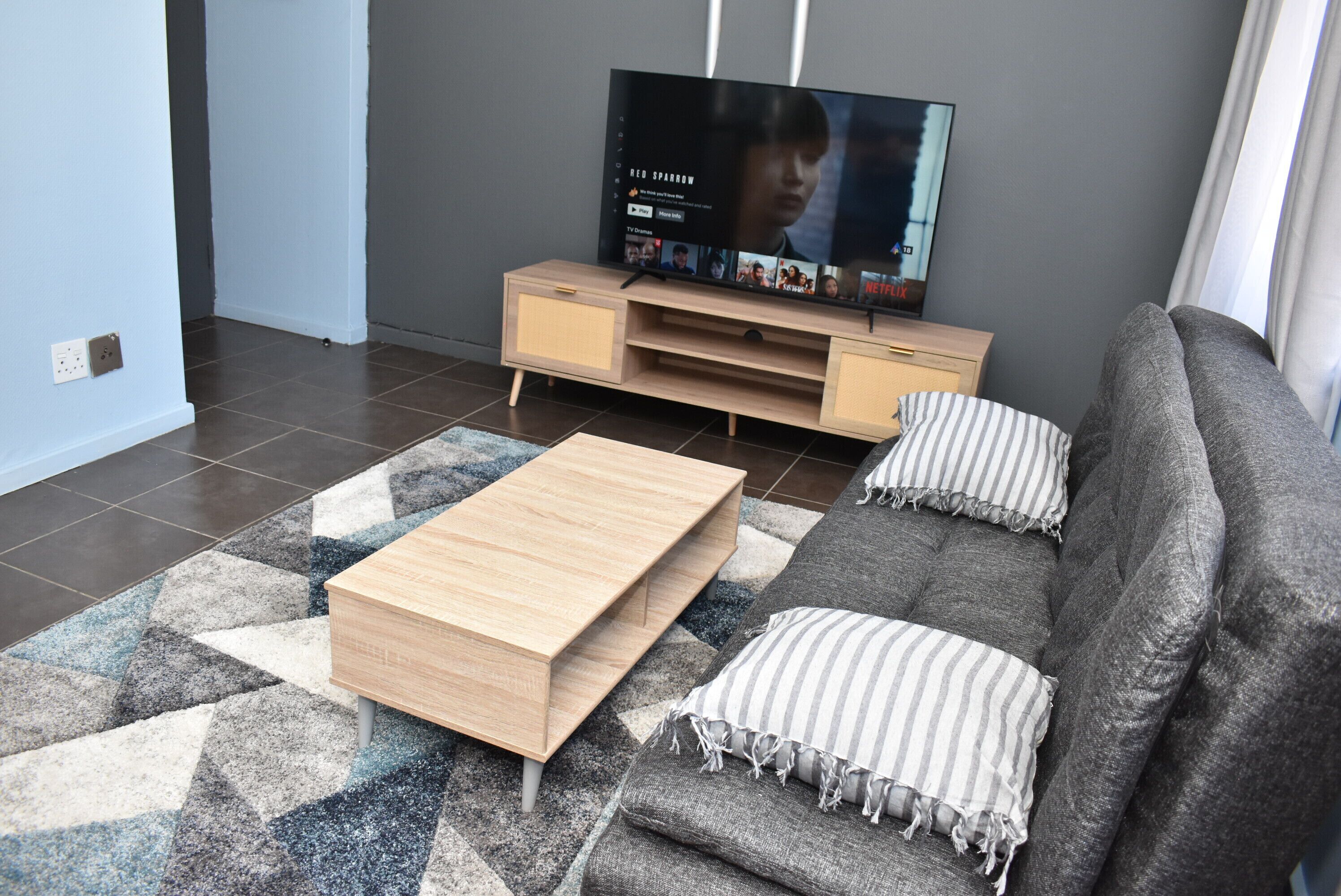 A modern living room with a flat-screen TV, a wooden coffee table, a grey sofa, and striped pillows.