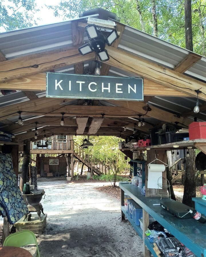 Een landelijke buitenkeuken met een bordje met de tekst 'KITCHEN', houten balken, hangende lampen en diverse kookgerei en bakspullen.