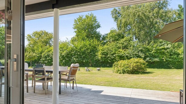Puisesta laudasta rakennettu terassi, jossa on pöytä ja tuoleja, pergola ja näkymä vehreälle, puita kasvavalle alueelle.