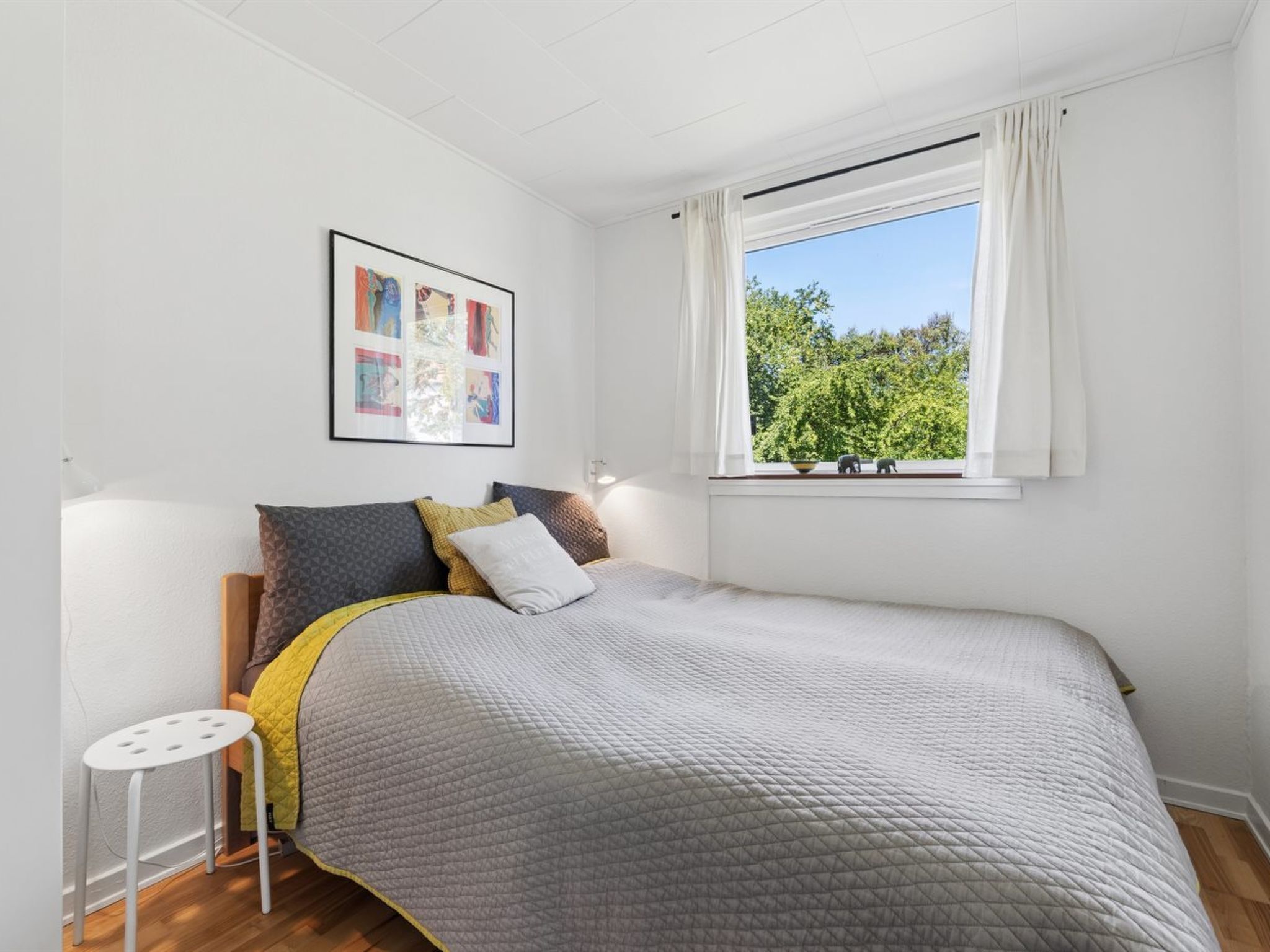 A bedroom with a bed, a bedside table, a window with curtains, and a framed picture on the wall.