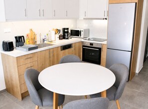Uma cozinha moderna com armários brancos, forno embutido, micro-ondas e máquina de café. Há uma mesa de jantar redonda com quatro cadeiras cinzentas.
