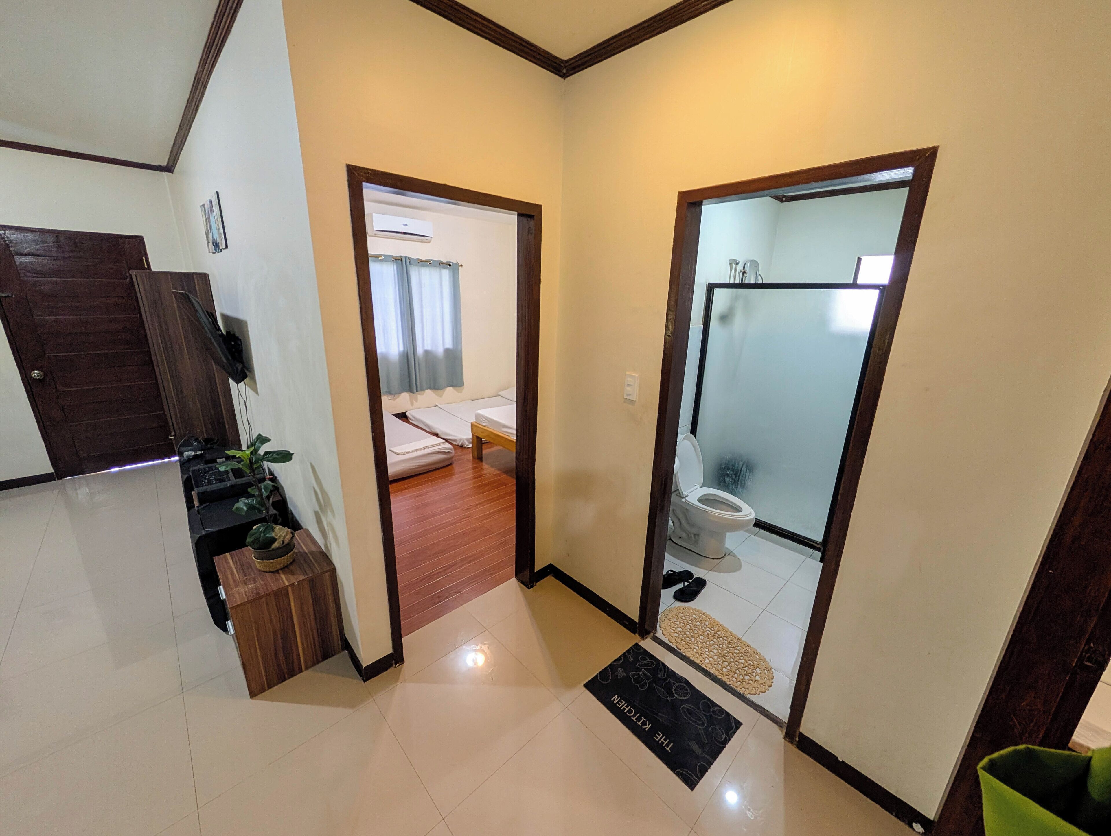 A hotel room with a wooden door, a small desk with a plant, a bed with white bedding, and a bathroom with a toilet and shower.