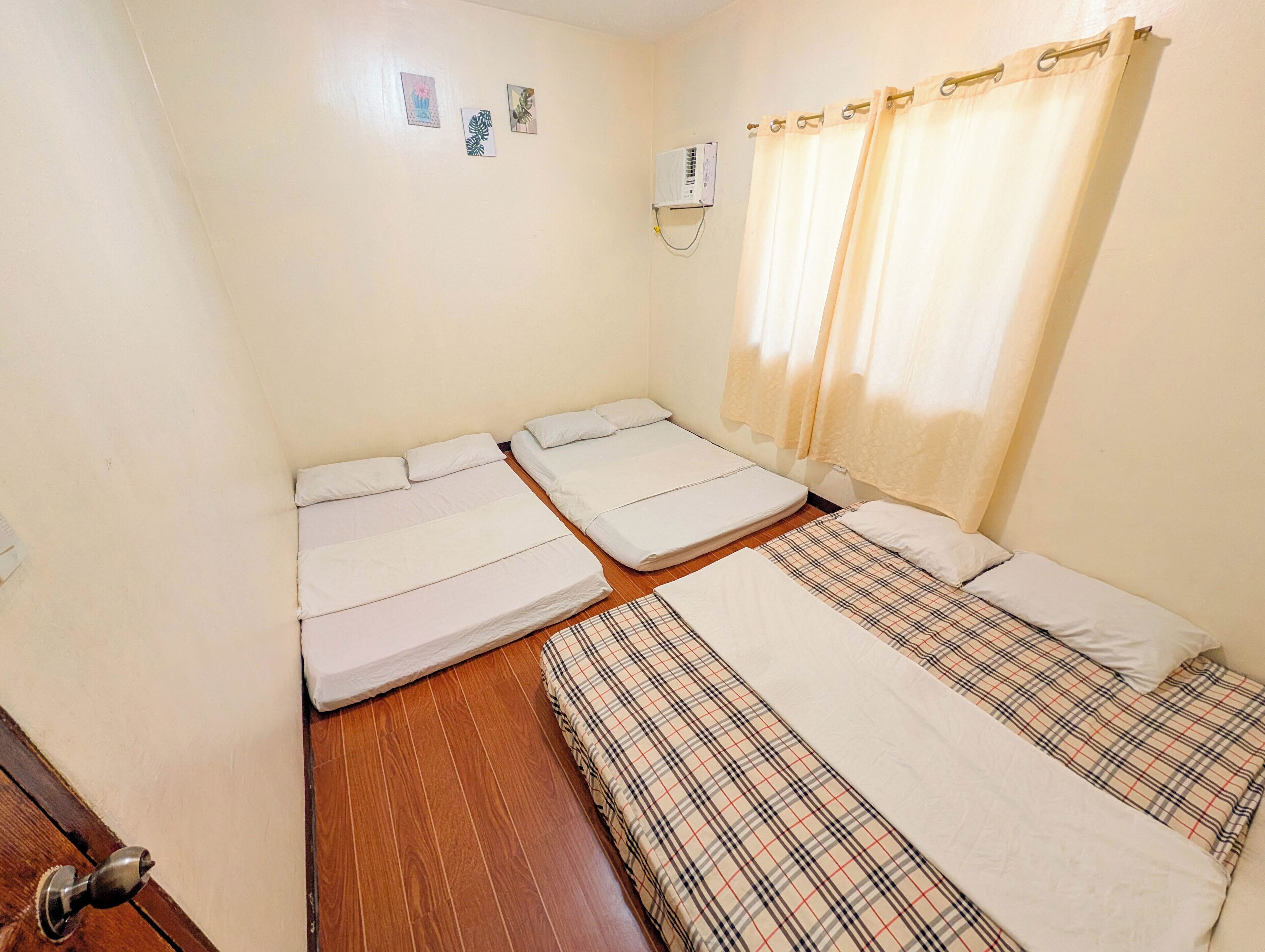 Une chambre avec deux lits, un plancher en bois, un climatiseur fixé au mur et une fenêtre avec des rideaux.