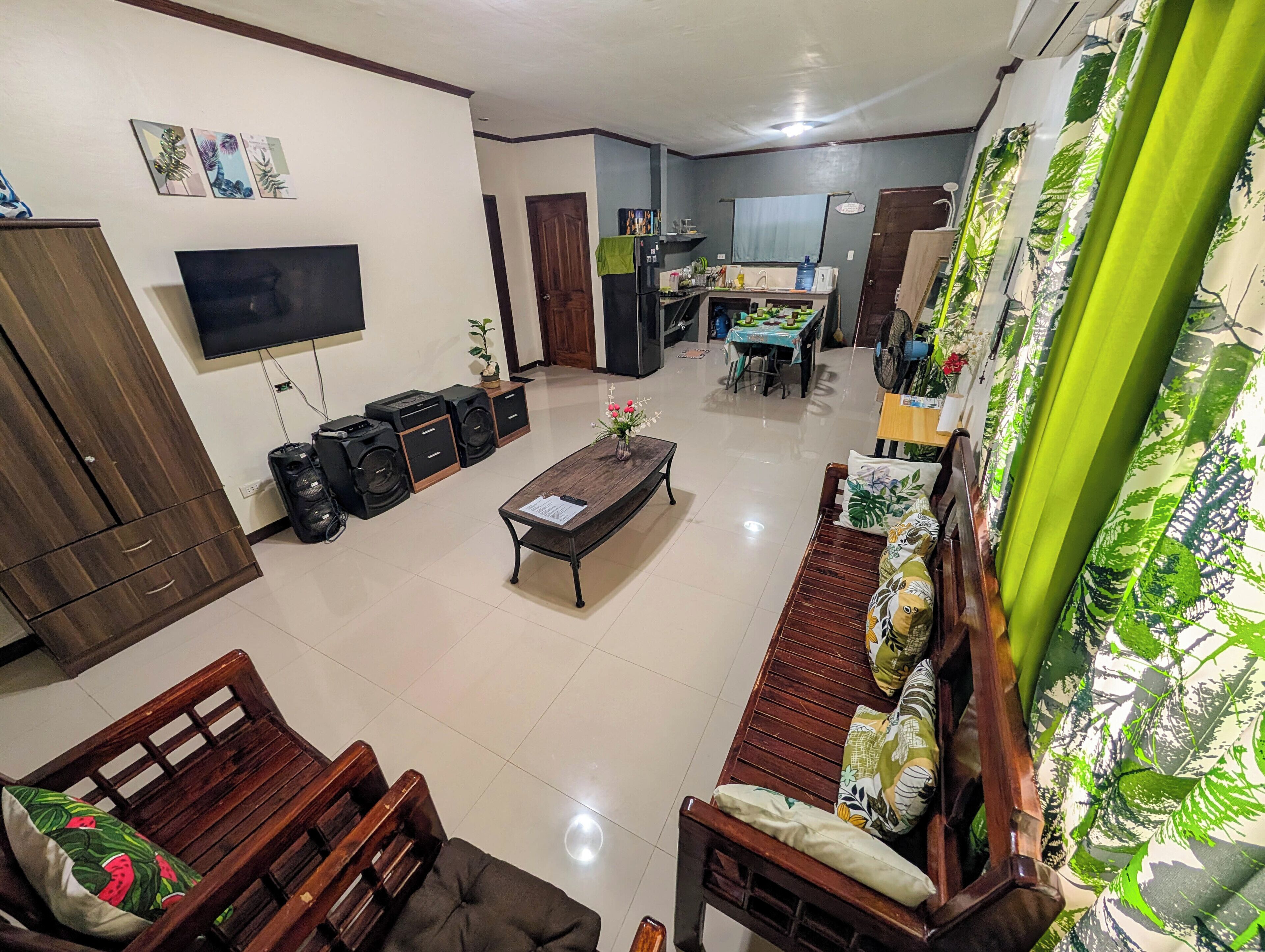 A living room with a sofa, a coffee table, and a TV. There is a kitchen in the background with a dining table and chairs.
