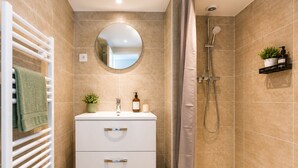 A bathroom with a white vanity, a round mirror, a shower with a handheld showerhead, a towel rack, a small plant, and a trash bin. - Charming 1-bedroom apartment near Geneva, between the lake and the mountains (Bons-en-Chablais)