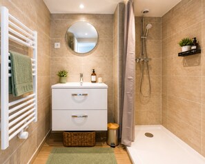 A bathroom with a white vanity, a round mirror, a shower with a handheld showerhead, a towel rack, a small plant, and a trash bin. - Charming 1-bedroom apartment near Geneva, between the lake and the mountains (Bons-en-Chablais)