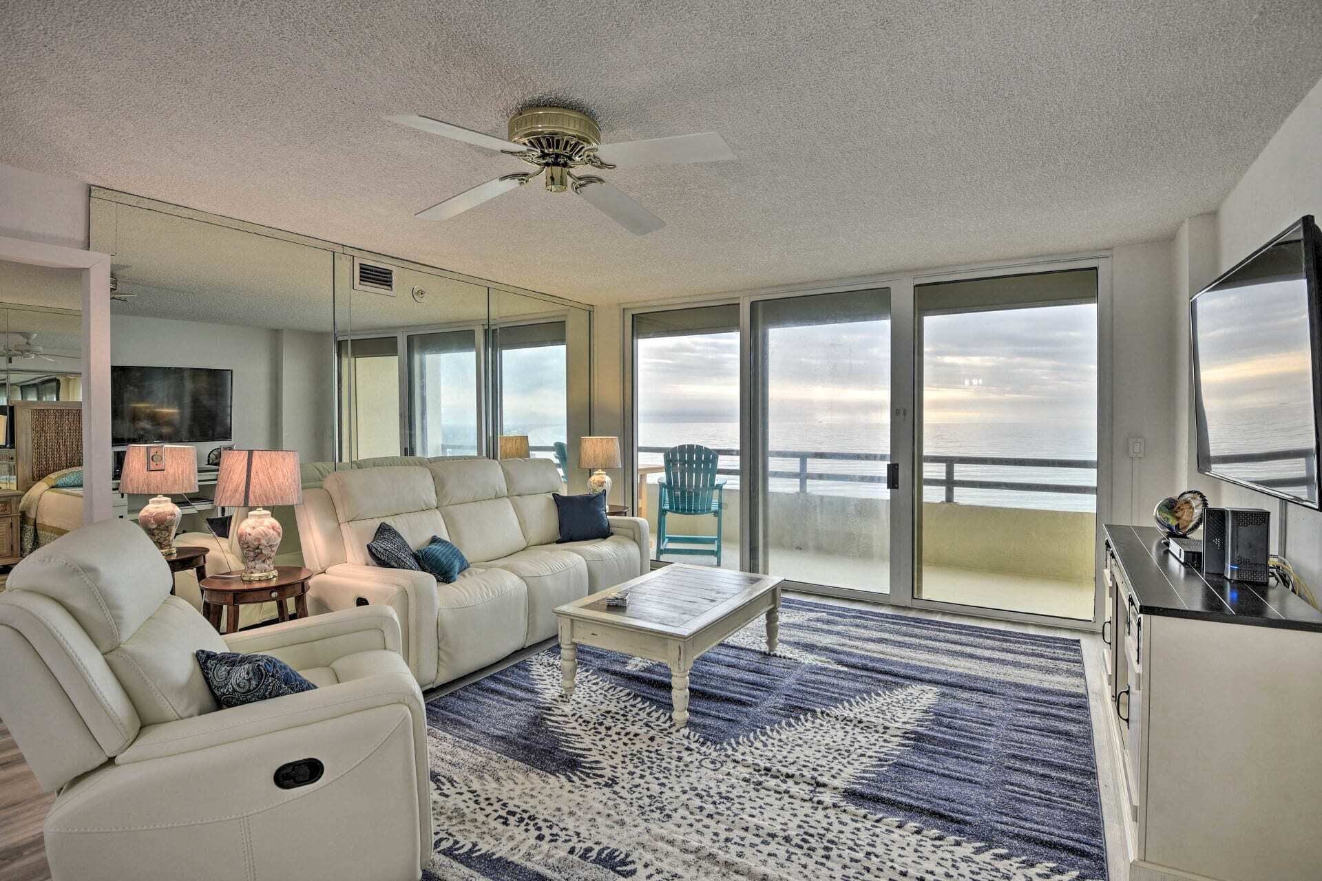 A spacious living room with a large window, a ceiling fan, and a view of the ocean.