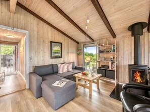 Ein gemütliches Zimmer mit Holzbalkendecke, Kamin, einer Couch, einem Tisch mit Obstschale und einem Bücherregal.