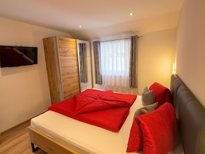 Ein Schlafzimmer mit einem Bett, einem an der Wand montierten Fernseher, einem Kleiderschrank und einem Fenster mit Vorhängen.