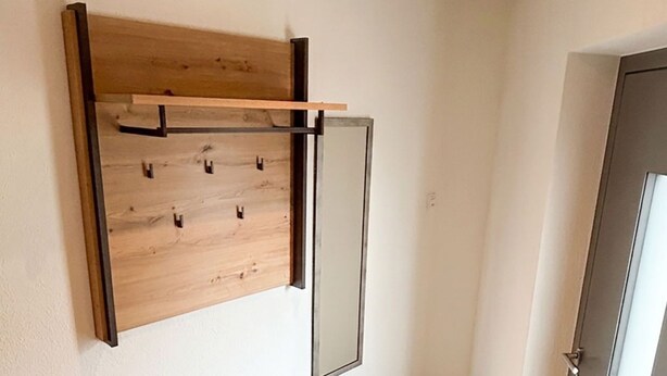 Minimalistinen huone, jossa on puinen seinään kiinnitetty naulakko, laatikoilla varustettu penkki ja lasipaneelilla varustettu ovi.