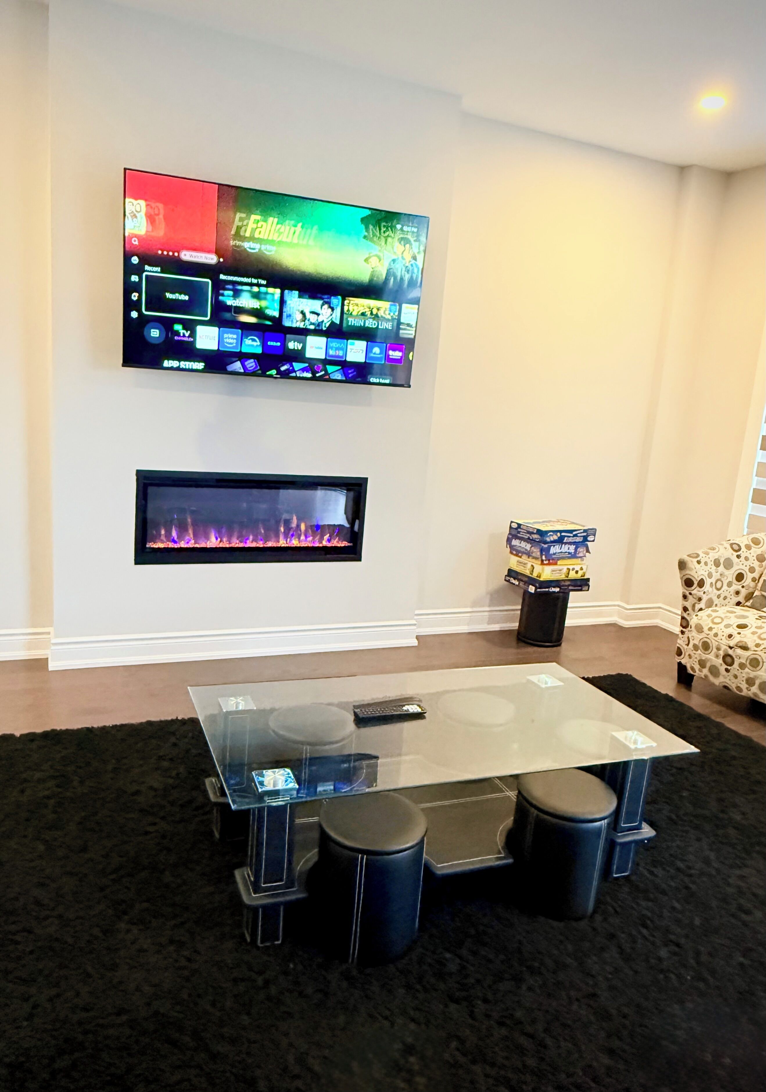 A living room with a flat-screen TV mounted on the wall, a fireplace below it, and a glass coffee table with stools.