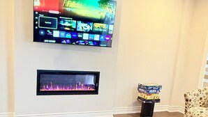 Sala de estar com televisão de tela plana na parede, lareira abaixo dela e mesa de centro de vidro com bancos.
