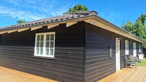 Una cabaña de madera con fachada oscura, una ventana de marco blanco y un área de terraza con mobiliario exterior.