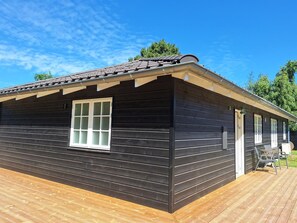 Eine Holzhütte mit dunkler Fassade, einem Fenster mit weißem Rahmen und einer Terrassenfläche mit Gartenmöbeln.