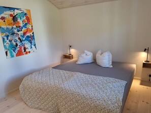 Une chambre à coucher avec un lit, deux tables de chevet, une lampe fixée au mur et un tableau abstrait aux couleurs vives.
