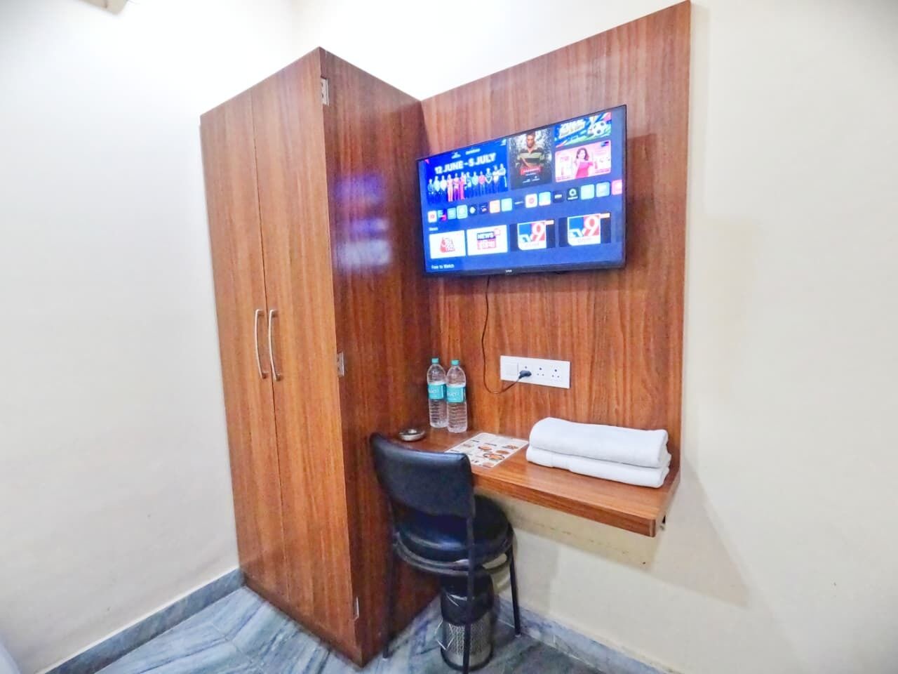 A small room with a wooden wardrobe, a desk with a chair, a flat-screen TV mounted on the wall, and a small shelf with a towel.