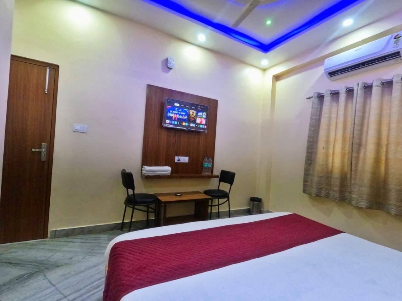 Habitación de hotel con una cama, un televisor montado en la pared, una mesita con dos sillas y una cortina en la ventana.