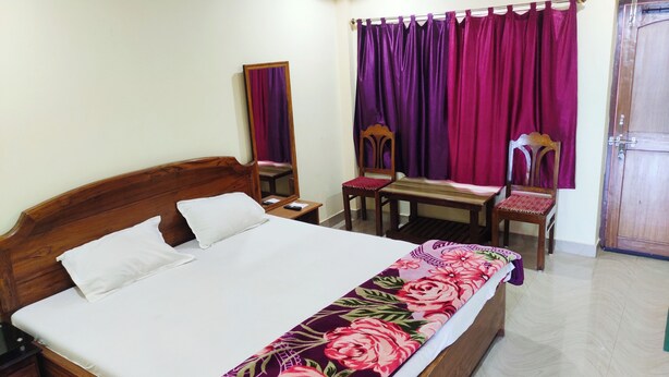 A bedroom with a bed, a wooden nightstand, a chair, a table, a mirror, and a door. - LUXURY INN  (Chakradharpur)