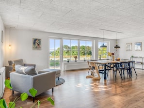 En rummelig opholdsstue med sofa, spisebord og stole, store vinduer med udsigt til grønne områder, og et loft med et gittermønster.