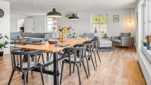 Uma sala de estar ampla com uma mesa de jantar, cadeiras e um sofá. Cozinha visível ao fundo, com uma janela.