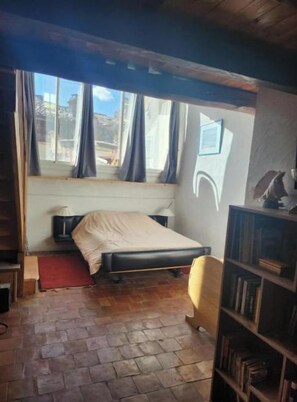 A bedroom with a bed, a nightstand, a bookshelf, and a window with curtains. - The Boat on the Rooftops (Cotignac)