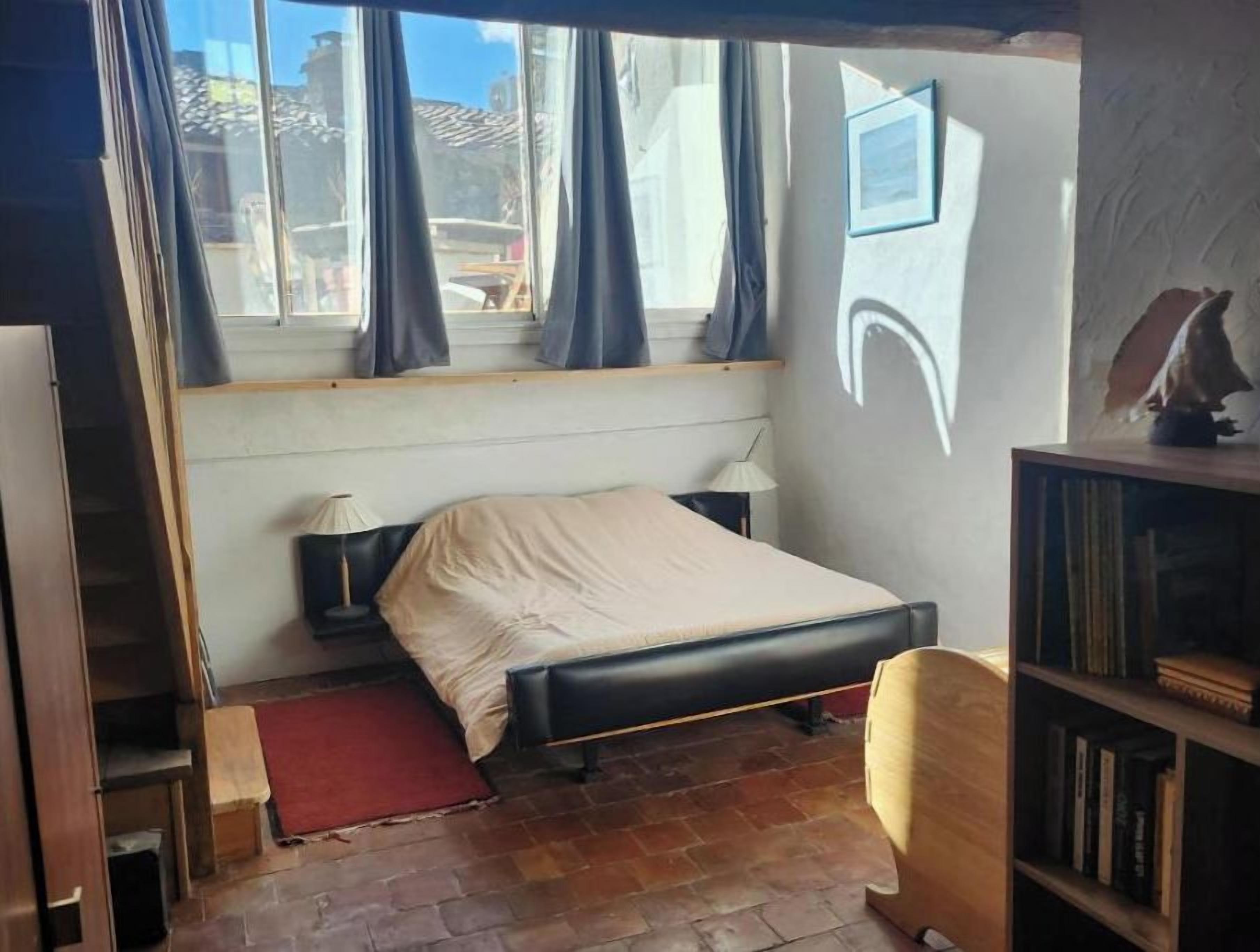 Un dormitori amb un llit, una tauleta de nit, una làmpada, una biblioteca i una finestra amb cortines.