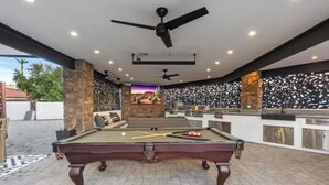 Una mesa de billar, un ventilador de techo, una zona de estar con vistas al exterior y una barra con un muro decorativo.